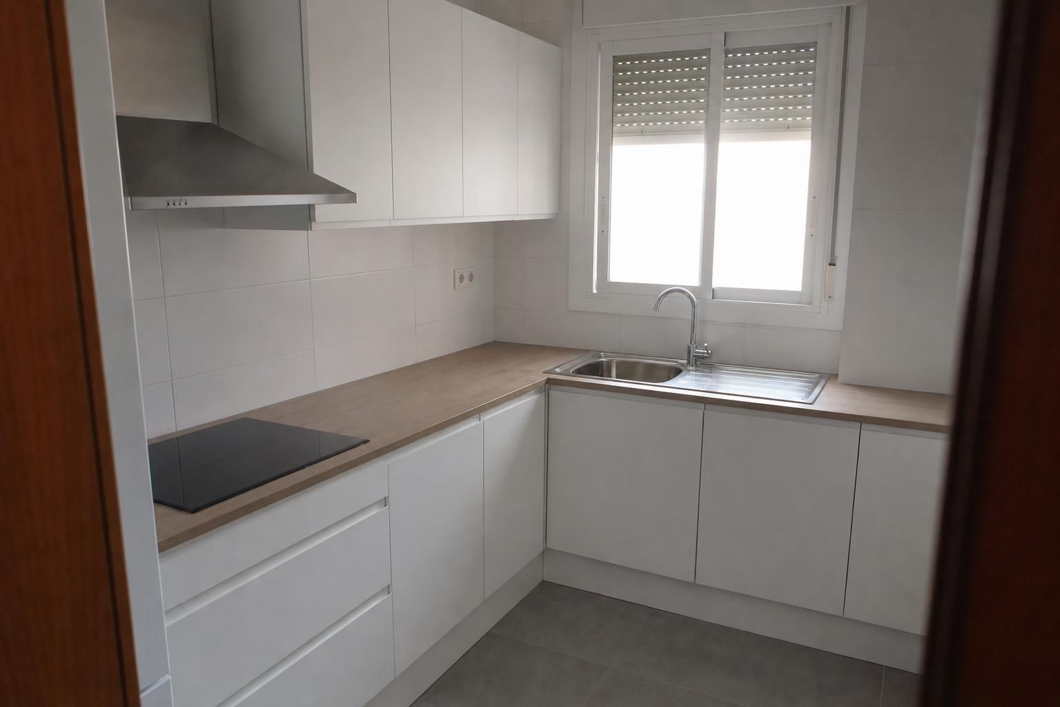 Cocina reformada en L con muebles blancos, encimera clara y fregadero bajo la ventana